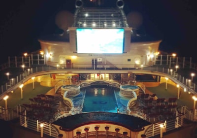 A luxury cruise ship deck featuring a pool surrounded by lounge chairs and warm ambient lighting. A large screen overlooks the area, showing an image against a dark night sky.