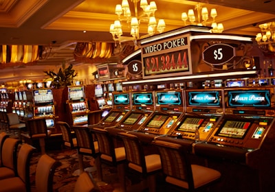 A casino floor featuring rows of brightly lit video poker machines. The area is illuminated by elegant chandeliers, with a display board showing a large sum of money, suggesting a possible jackpot. The rich decor includes luxurious carpets and comfortable seating, adding to the opulent atmosphere.