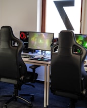 A row of gaming stations equipped with large monitors, gaming chairs, keyboards, and headphones. The setup is organized and ready for gaming, featuring a colorful screen display. The room has a modern and tech-savvy ambiance.
