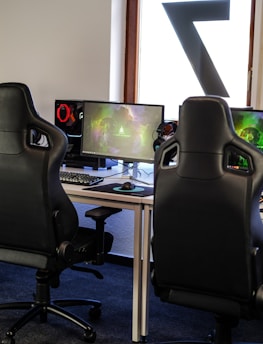 A row of gaming stations equipped with large monitors, gaming chairs, keyboards, and headphones. The setup is organized and ready for gaming, featuring a colorful screen display. The room has a modern and tech-savvy ambiance.