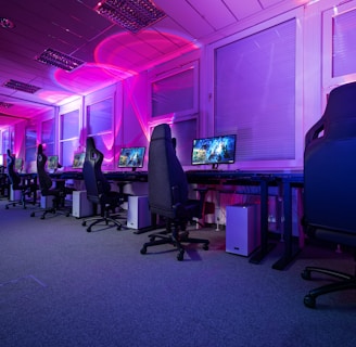 A row of gaming computers with large monitors and racing-style chairs is set up along the interior of a modern room. The space is illuminated with vibrant purple and blue LED lighting, creating a futuristic atmosphere. The room is organized and clean, suggesting a professional gaming or esports environment.