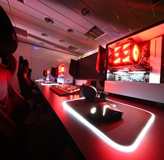 A modern gaming setup featuring several high-end computers with illuminated red interior lighting. The room is equipped with gaming chairs exhibiting a sleek black and red design. Multiple monitors are arranged on a long desk, and peripheral devices like keyboards and mice are uniformly placed, reflecting an ambiance ideal for gaming or competitive play.