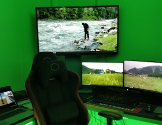 A gaming setup with a large curved monitor showing a scenic image of a river and forest. Several smaller monitors display nature scenes, and a laptop is open showing a different video. A comfortable gaming chair is positioned in front of the setup. The room is illuminated by green lighting, and the desktop tower has colorful RGB fans.