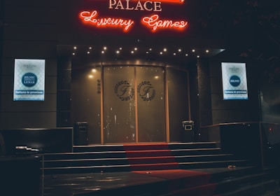 A neon-lit casino entrance with a red carpet leading up to large double doors. The sign reads 'Fortuna Palace Luxury Games' in bright red and white neon lights. The dimly lit atmosphere suggests a night-time setting. There are two digital displays on either side of the entrance showing promotional content.